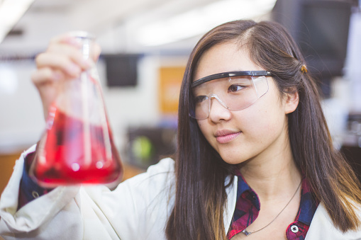 An ethnic high school girl works in the chemistry laboratory. She is wearing safety glasses and is holding up a beaker filled with liquid and closely examining it.