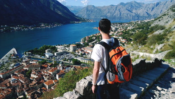 Young teen admires the view during the ascent to the fortress in Kotor in Montenegro. Male brunette in white t-shirt and bright orange backpack stock photo