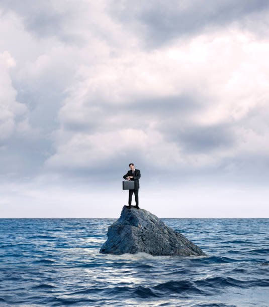 Businessman Marooned On Rock In Ocean Checks His Watch A businessman holding a briefcase stands on a rock in the middle of the ocean as he checks his wristwatch for the time. stranded stock pictures, royalty-free photos & images
