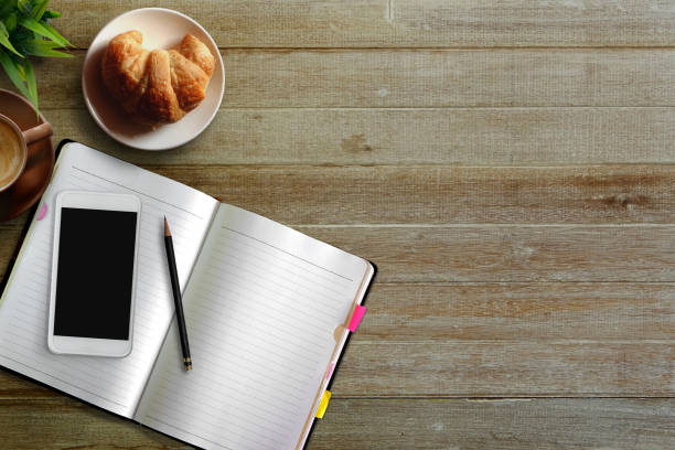 White porcelain cup of coffee, tasty chololate muffin, pieces of black chocolate, notebook, classic pen and a smartphone on a working table. Business day concept.