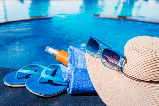 Blue slippers with sunscreen cream, towel, straw hat and sunglasses on border of a swimming pool - holiday tropical concept