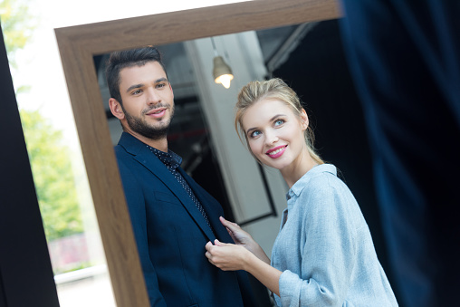 smiling young couple looking at mirror while choosing suit in boutique
