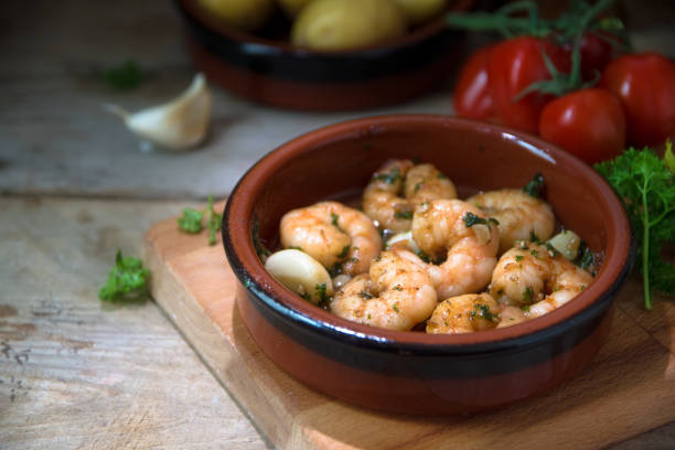 gamberi o gamberi in aglio e olio d'oliva in una ciotola di tapas, un antipasto spagnolo su una tavola di legno, patate, pomodori ed erbe sullo sfondo, stile rustico scuro - ajillo foto e immagini stock