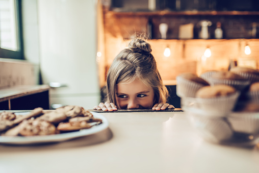 Charming little girl on kitchen is looking from under the table on sweets. Ready to eat some cookies and cakes.