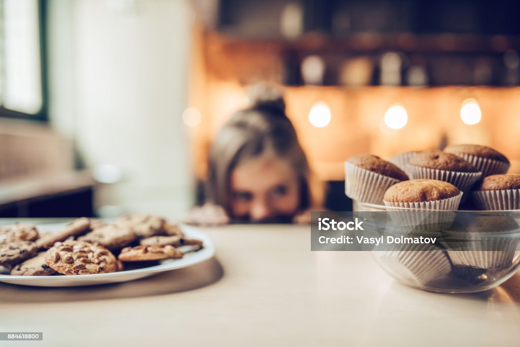 Little girl on kitchen. Charming little girl on kitchen is looking from under the table on sweets. Ready to eat some cookies and cakes. Craving Stock Photo