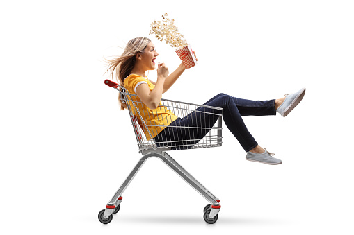 Young woman with a box of popcorn riding inside a shopping cart isolated on white background