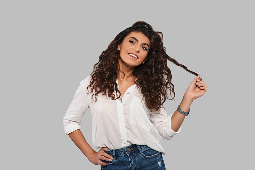 Attractive young woman playing with her hair and smiling while standing against grey background