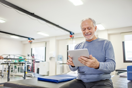 Senior man smiling while using digital tablet in rehab center. Elderly male is sitting on bed in health club. He is in sports clothing at gym.
