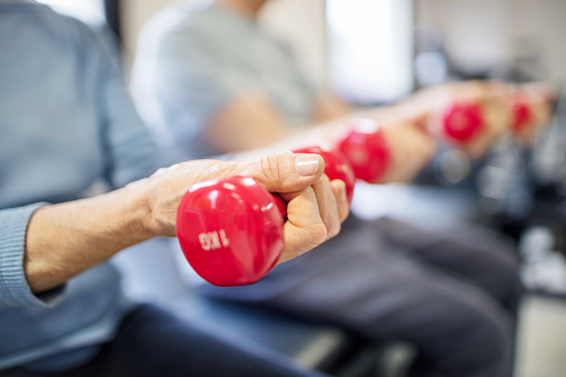 Midsection of senior woman lifting dumbbells in rehabilitation center. Elderly female is exercising with man in gym. Aged patient is in health club.