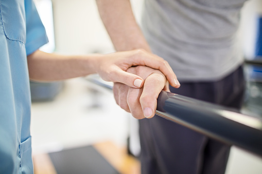 Cropped image of female nurse touching senior man's hand on railing. Medical professional is assisting senior man to walk. Elderly patient is walking between parallel bars in rehabilitation center.