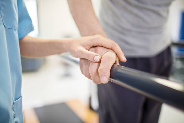 Female nurse touching senior man's hand on railing Cropped image of female nurse touching senior man's hand on railing. Medical professional is assisting senior man to walk. Elderly patient is walking between parallel bars in rehabilitation center. recovery stock pictures, royalty-free photos & images