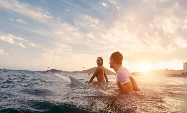 forma pareja surfeando en puesta del sol - amigos surfistas divirtiéndose dentro de océano - extremo deporte y vacaciones concepto - foco en la cabeza del hombre - original sol tonos de color - surf fotografías e imágenes de stock