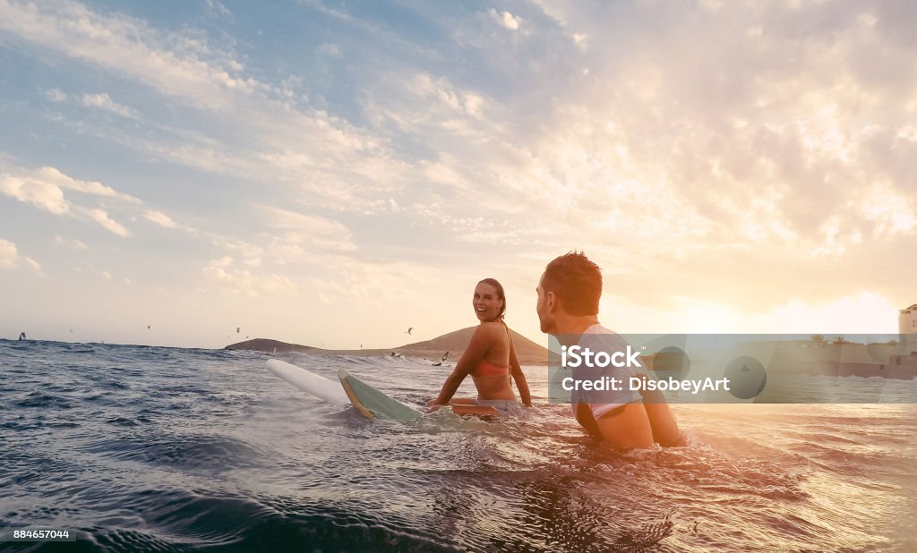 Fit couple surfing at sunset - Surfers friends having fun inside ocean - Extreme sport and vacation concept - Focus on man head - Original sun color tones Surfing Stock Photo