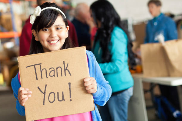 Adorable little girl holds thank you sign during food drive An adorable elementary age little girl stands in front of a group of bustling food bank volunteers and smiles for the camera as she holds a hand-written cardboard sign that says, "Thank you!" charitable foundation photos stock pictures, royalty-free photos & images