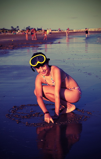 smiling little girl with diving mask draws a big heart in the sand of the shore in summer