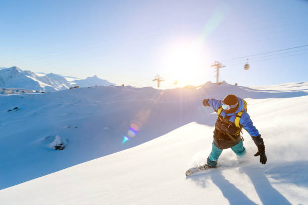 istock A snowboarder in a ski mask and a backpack is riding on a snow-covered slope leaving behind a snow powder against the blue sky and the setting sun 885102510