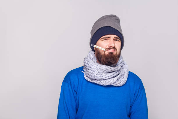 Young adult bearded man have temperature, holding thermometer in mouth. Young adult bearded man have temperature, holding thermometer in mouth. Studio shot, isolated on gray background sneezeweed stock pictures, royalty-free photos & images