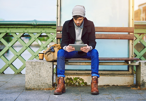 Shot of a young man using a digital tablet while waiting at a city bus stop