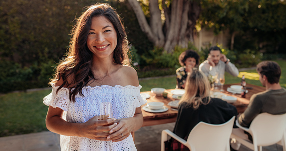 Beautiful asian woman standing outdoors with friends sitting in background having food. Woman with a drinks at outdoor party.