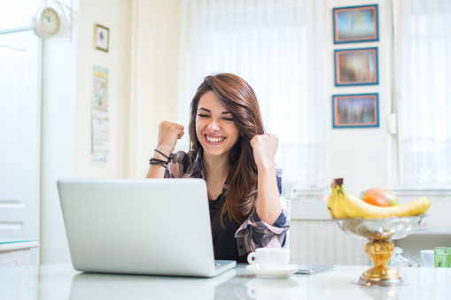 Portrait of happy young woman celebrating success with arms up in front of laptop at home.