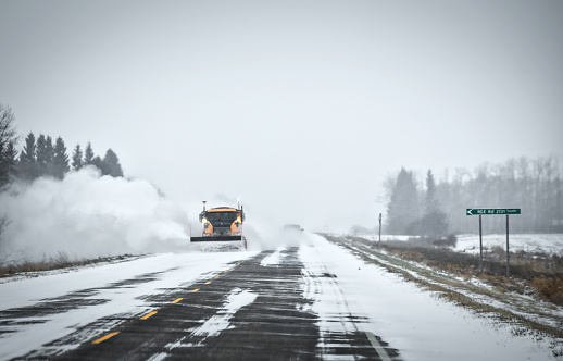 A snowplow vehicle driving along a highway clearing billowing clouds of snow off in a winter snow storm