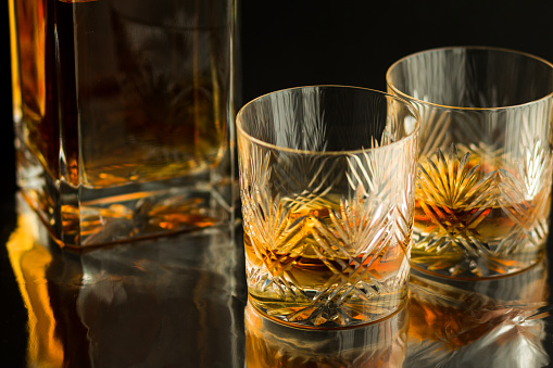 Close up color image depicting two fine crystal glasses filled with a measure of golden Scotch single malt whisky. The cut glass tumblers rest on a silver reflective bar counter surface, and slightly defocused in the background is a glass decanter filled with whisky. Room for copy space, isolated on black background.