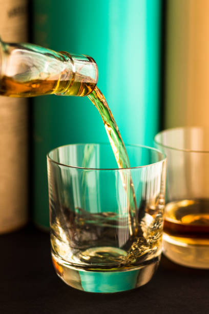 Whisky being poured from clear glass bottle into glass tumbler with Scotch malt whisky bottles in background