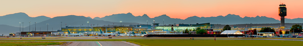 Panorama of a modern airport and white planes in electric light lights against the background of an orange evening sky and silhouettes of mountains