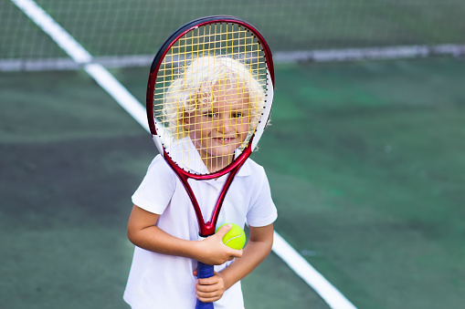 Child playing tennis on outdoor court. Little boy with tennis racket and ball in sport club. Active exercise for kids. Summer activities for children. Training for young kid. Child learning to play.