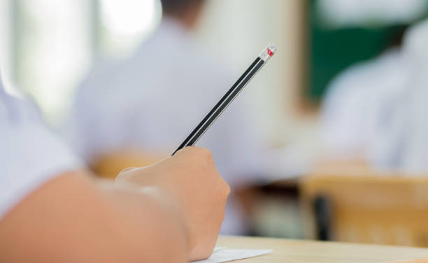 istock Hands with pencil over application form, Students taking exams, writing examination room with undergraduate students inside. Student sitting learning lessons doing final exam in classroom. 885708974