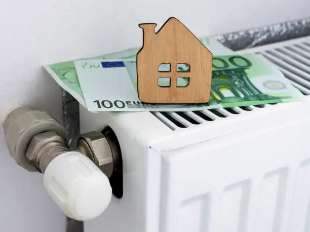 Photo of A small wooden house on the background of euro banknotes and radiator heating. Payment for heating in winter. Symbolic image, selective focus.