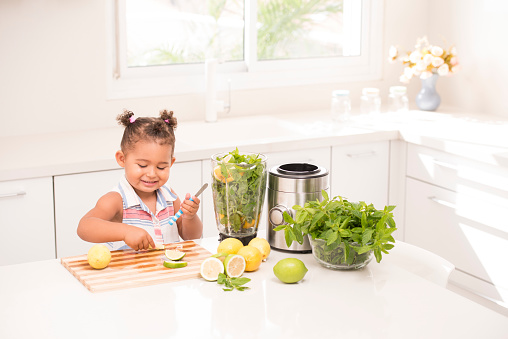 3 years old toddler girl on a sunny morning, cutting a natural fresh lemon and mixing it with mint leaves for making lemonade drink.