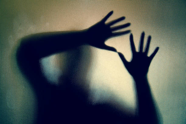 Spooky silhouette of woman with hands pressed against glass window Colour backlit image of the silhouette of a woman with her hands pressed against a glass window. The silhouette is distorted, and the arms elongated, giving an alien-like quality. The image is sinister and foreboding, with an element of horror. It is as if the 'woman' is trying to escape from behind the glass. Horizontal image with copy space. Trafficking stock pictures, royalty-free photos & images