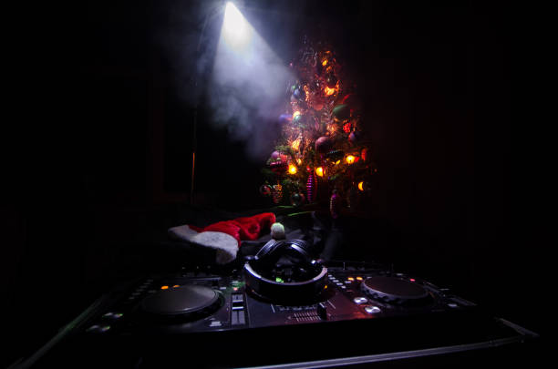 Dj mixer with headphones on dark nightclub background with Christmas tree New Year Eve. Close up view of New Year elements or symbols (Santa Clause, Snowman, Dog 2018, gift box) on a Dj table. mezclador de dj con los auriculares en el fondo oscuro de la discoteca con árbol de navidad la víspera de año nuevo. cerrar vista de año nuevo os o símbolos (santa claus, muñeco de nieve, perro 2018, caja de regalo) en una mesa de dj. tonos
