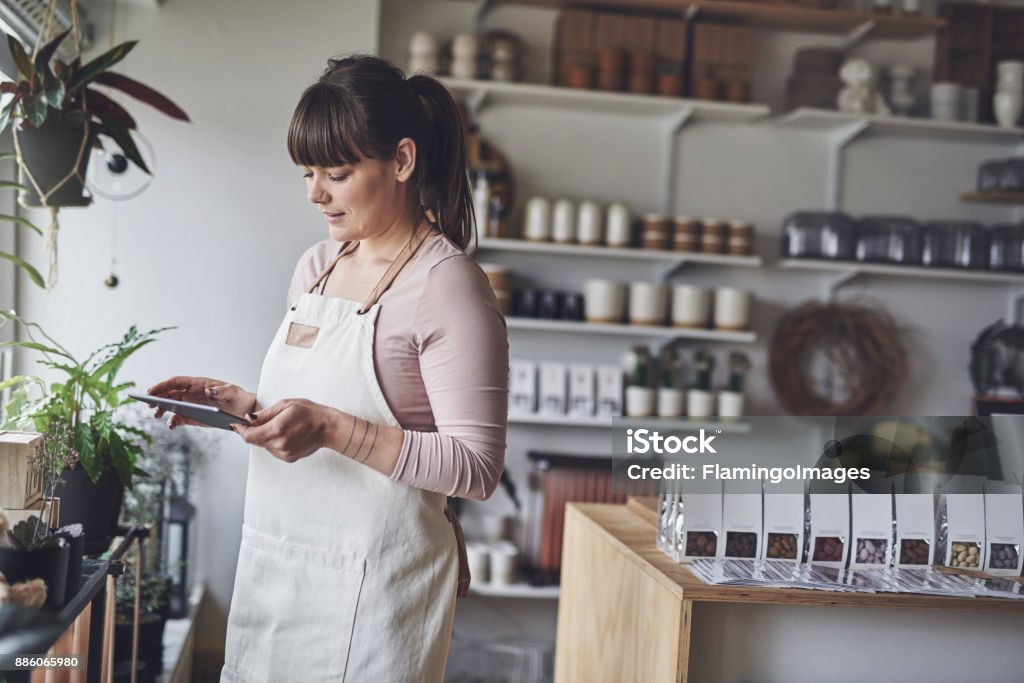 Laughing florist arranging a floral display in her shop Young female florist checking her online orders with a digital tablet while standing in her flower shop Small Business Stock Photo