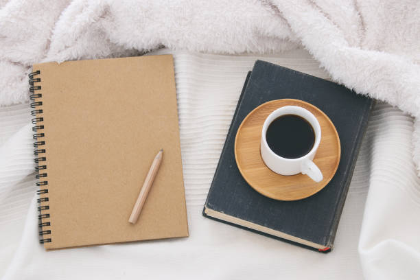 istock Cup of coffee on the old book next to empty notebook over cozy and white blanket. Top view. 886098154