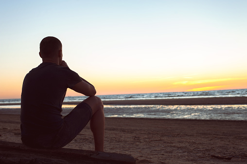 Burdened by unseen worries. Sad lonely man sits on wooden log at seashore looking forward. Vie from backside. Multicolored sunset background. Horizontal summertime outdoors image slightly filtered.
