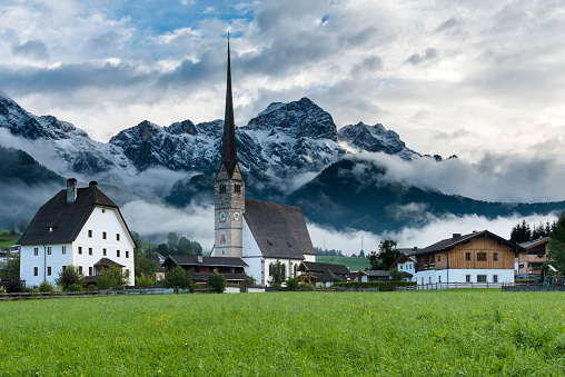 Maria Alm, Austria steeple shot from pasture in foreground with low hanging clouds and snow capped mountains in the background.