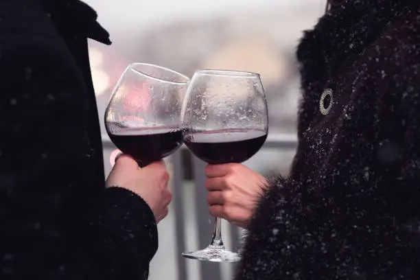 Crop shot of couple in outerwear clinking with wineglasses standing in snowflakes.