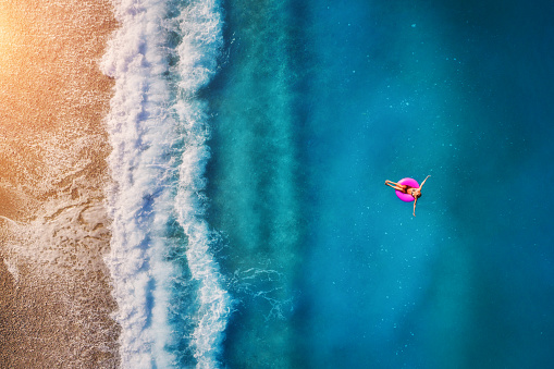 Aerial view of young woman swimming on the pink swim ring in the transparent turquoise sea in Oludeniz. Summer seascape with girl, beach, beautiful waves, blue water at sunset. Top view from drone