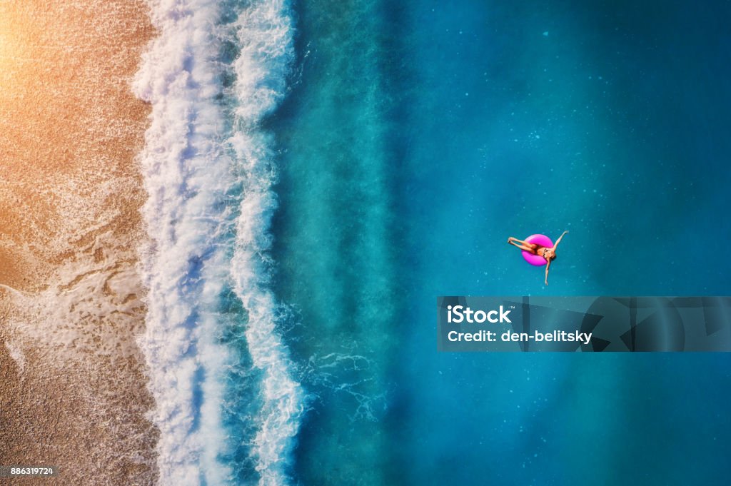 Aerial view of young woman swimming on the pink swim ring in the transparent turquoise sea in Oludeniz. Summer seascape with girl, beach, beautiful waves, blue water at sunset. Top view from drone Beach Stock Photo