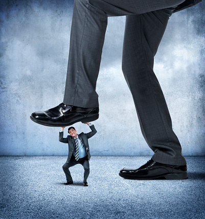 A small businessman looks up as he is about to get stepped on by a very large businessman.