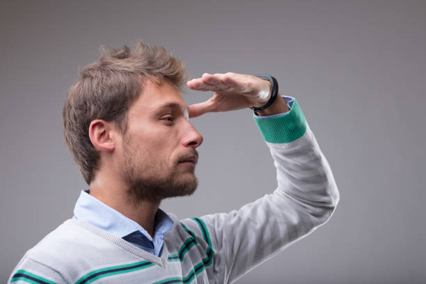 Blond man peering into the distance with his hand raised to his forehead in a closeup profile view over grey
