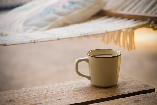 A cup of coffee mug on rustic wood table , blurred hammock on sand beach background. Coffee for relaxation