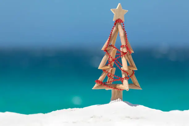 Summer sun shines down on a driftwood Christmas tree decorated with a string of red baubles on a beautiful white sandy beach with ocean background blur and copy space