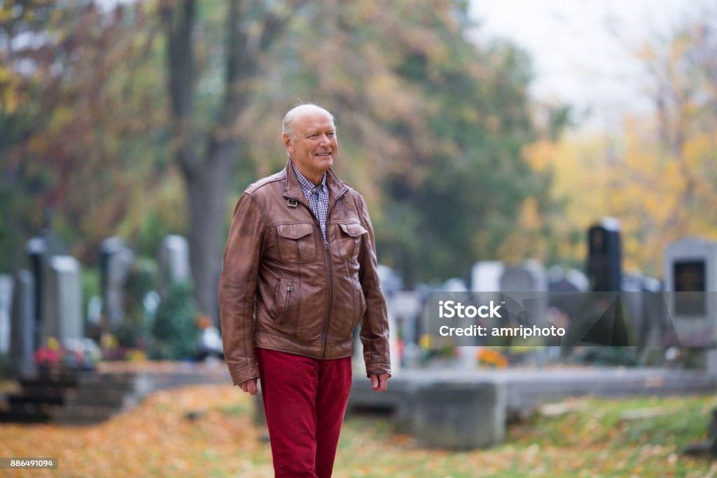 Seniors - 73 years active modern senior man taking a walk through cemetery in autumn senior man in graveyard series, all saints day cemetery with blurred tombs in the background shallow focus Public Park Stock Photo