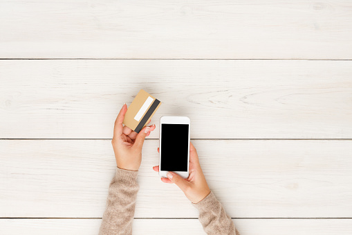 Online shopping in internet concept. Woman using credit card and blank smartphone, white wooden table background, top view