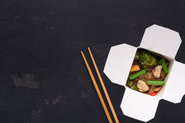 Asian food in delivery box on black background Asian restaurant food delivery. Funchoza with meat and vegetables, soy sauce and chilli pepper in white take away box and hashi on balck background, top view, copy space chinese takeout stock pictures, royalty-free photos & images