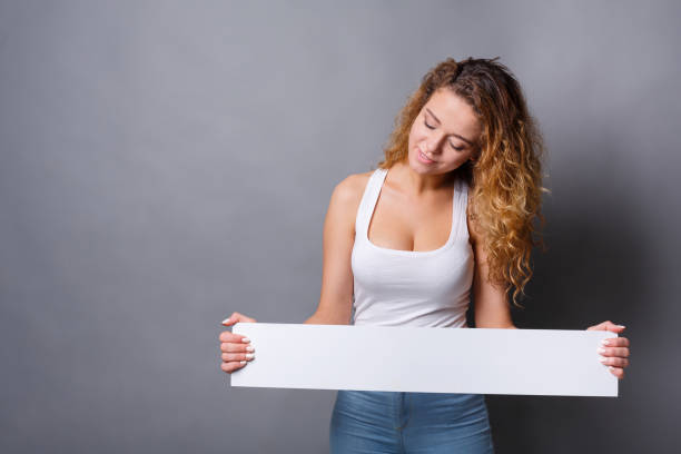 Young redhead woman holding blank white banner with copy space and showing on it, on gray background