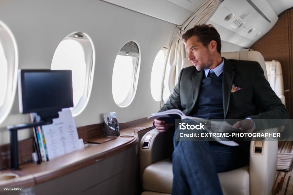 Man in private jet airplane Man sitting inside private jet airplane and looking outside the window while reading a magazine. Wealth Stock Photo